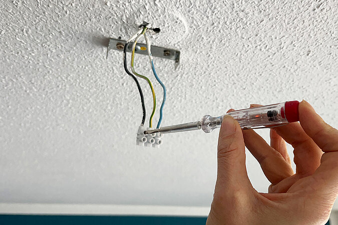 Die aus der Decke kommenden Stromkabel werden mit einem Spannungsprüfer auf Stromfluss überprüft. Der Stromprüfer zeigt vorhandenen Stromfluss an.
