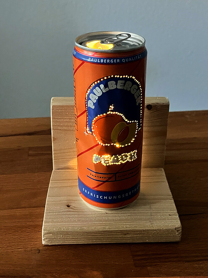 Leuchtende DIY-Tischampe aus gelochter Paulberger-Peach-Getränkedose auf einem L-förmigen Holzpodest in dunkler Umgebung.