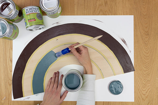 Wandleuchte im Regenbogen-Design bauen – Hand malt den zweiten Bogen in Blau an, um mehrfarbigen Effekt der Wandleuchte zu erzeugen.