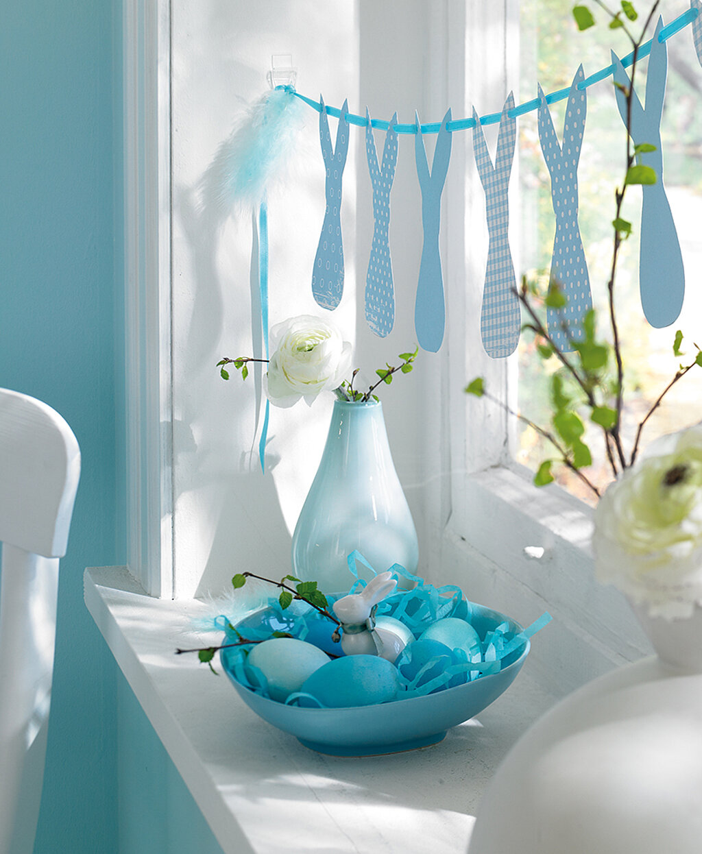 Fensterdeko mit blauer Hasen-Girlande und Feder am Band, auf der Fensterbank steht eine Schale mit blauen Eiern und eine Vase, Zweige ragen rechts ins Bild