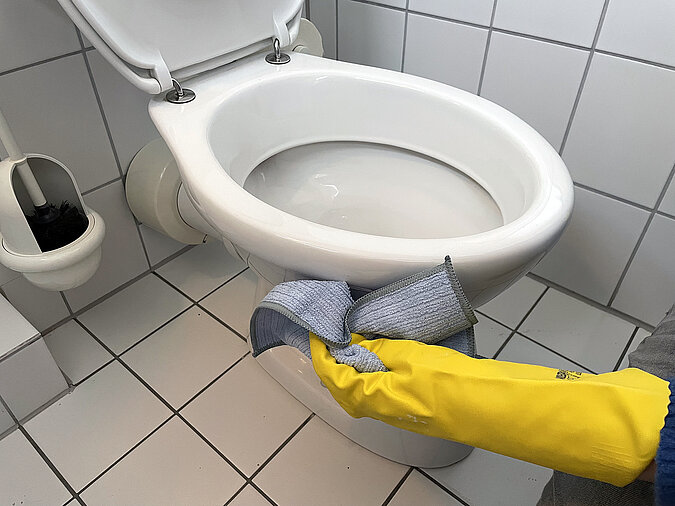 Hand mit Putzhandschuh wischt Toilette von aussen mit einem Lappen 