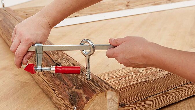 Mit Hilfe einer Schraubzwinge, der als Hebel eingesetzt wird, wird eine große Ösenschraube in den Holzbalken gedreht 