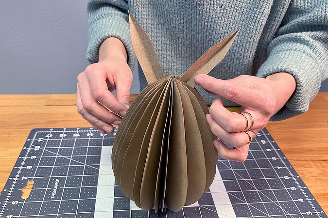 Hände stecken zwei lange Kartonohren oben in den gefalteten 3D-Körper und zeigen die Position