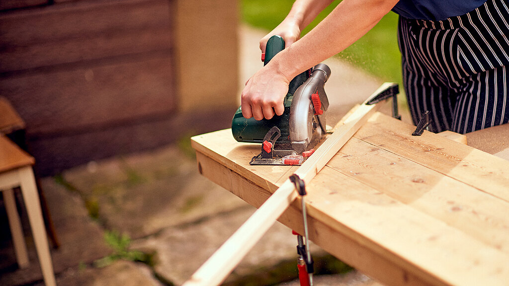 Eckbank Küche bauen – Holzbrett wird mit einer Handkreissäge auf der Werkbank zugeschnitten, um Bauteile für die Küchen-Eckbank vorzubereiten