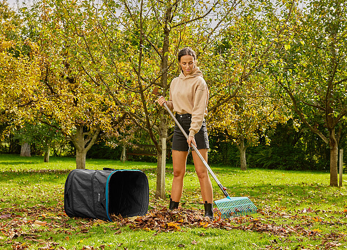 junge sportliche Frau recht im Herbstgarten Laub zusammen, neben ihr ein Behaelter zum Laubsammeln