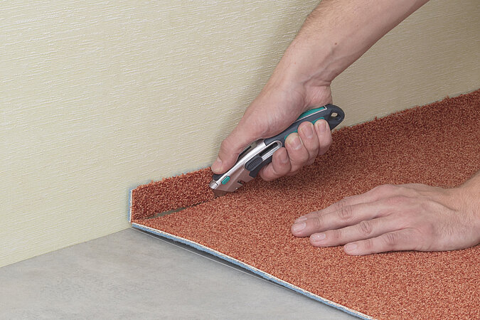 eine Hand schneidet mit Teppichmesser Teppich an der Wand zu