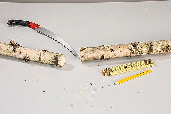 Birkenstämme für einen Raumteiler aus Birkenstämmen liegen mit Holzbohrer, Schrauben und Bleistift auf einer Arbeitsfläche