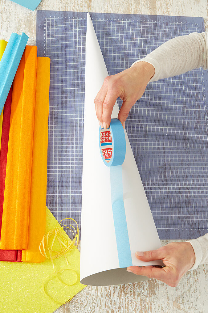 Auf einen weißen Schultüten-Rohling werden blaue Malerkrepp-Streifen für den Raketen-Look geklebt