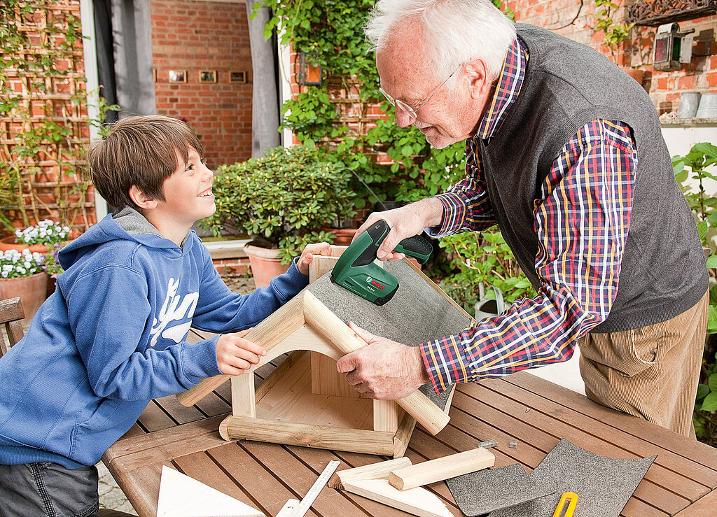 Tipps zum Heimwerken mit Kindern: Großvater und Enkel bauen gemeinsam ein Vogelhaus mit einem Elektrotacker – generationsübergreifendes DIY-Projekt im Garten.