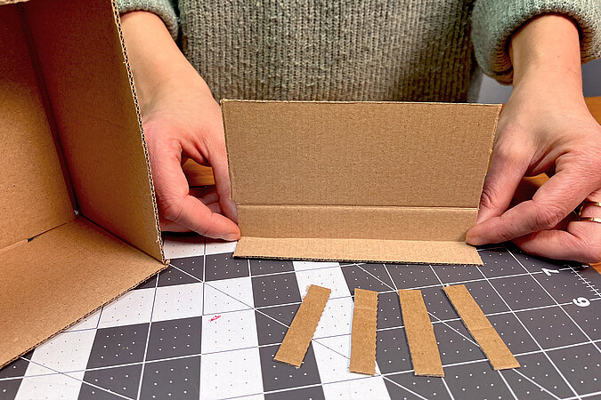 Um eine Trennwand in einen Karton einzubauen, wird ein Stück Pappe mit einem abgeknickten Stück und 4 Pappstreifen zum Fixieren gebraucht