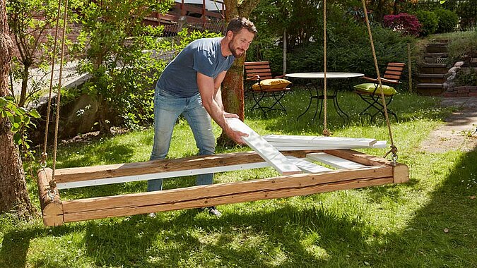 Um ein Gartenbett aus Holz aufzuhängen, bindet ein Mann Seile um einen stabilen Ast