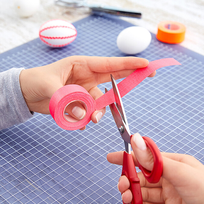 Hände schneiden einen Streifen pinkes Tape mit einer roten Schere auf der Schneidematte, im Hintergrund liegen Eier und eine orange Tape-Rolle