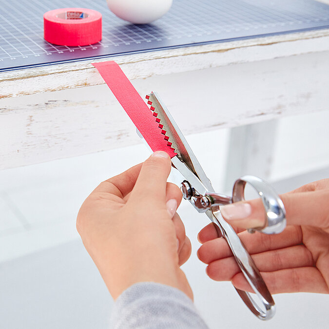 Hände schneiden pinkes Tape mit einer Zackenschere in Zickzackform, das Tape hängt über der Tischkante, Tape-Rolle und Ei liegen oben auf der Schneidematte