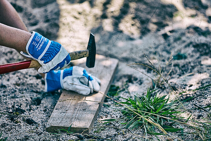 Tipps zum Heimwerken mit Kindern: Kinderhand mit Arbeitshandschuhen schlägt vorsichtig einen Nagel in ein Holzbrett – sicheres Üben handwerklicher Fähigkeiten im Freien.