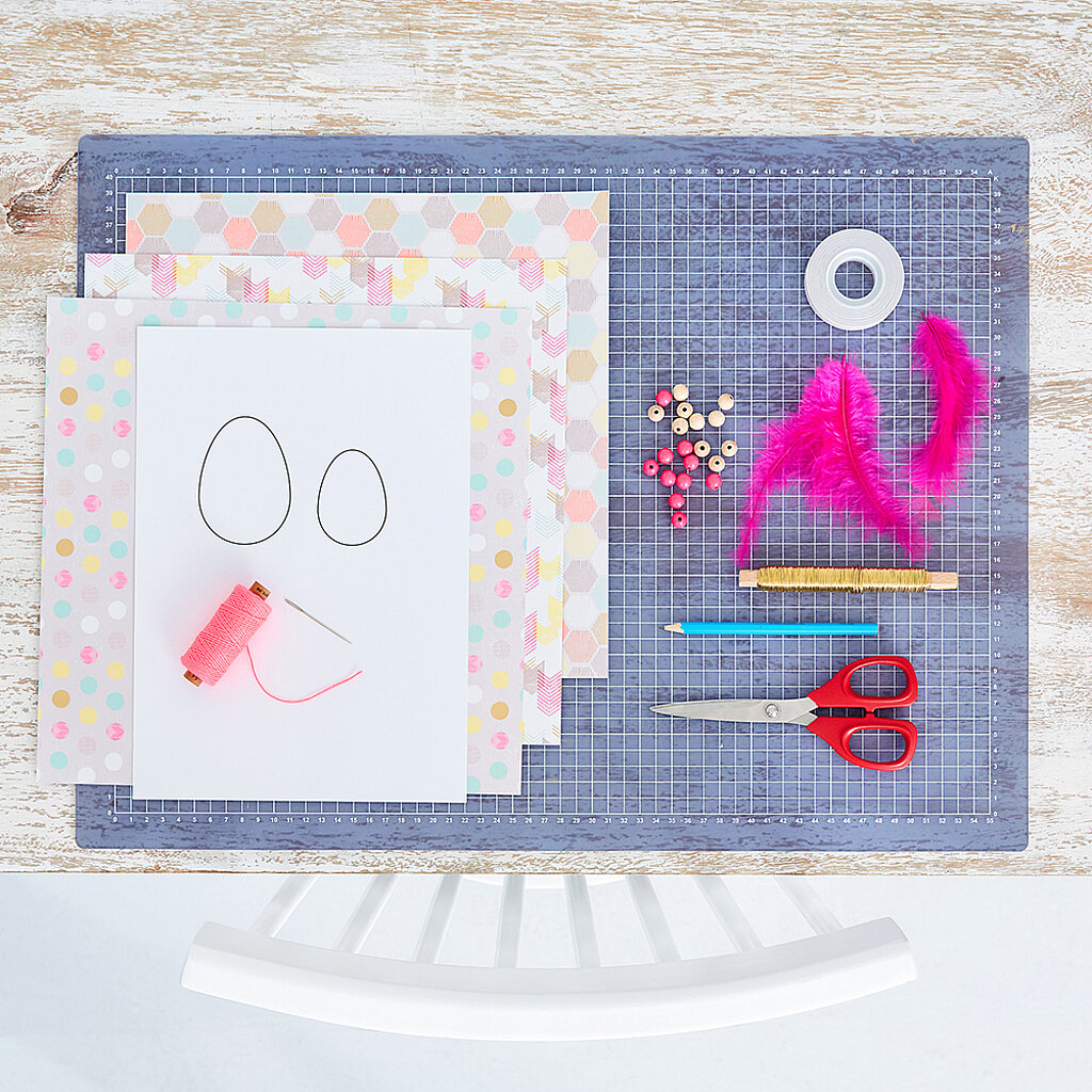 Materialübersicht für Ei-Anhänger mit gemustertem Bastelpapier, Schablone mit zwei Ei-Umrissen, pinkem Garn, Nadel, Draht, Holzperlen, pinken Federn, Klebeband, Bleistift und roter Schere auf Schneidematte