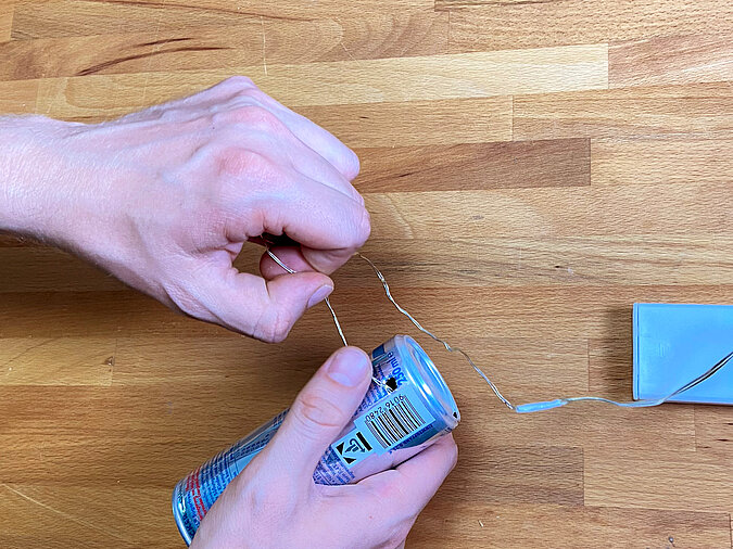Lichterkette wird durch das geprickelte Loch einer Getränkedose eingefädelt, daneben liegt das Batteriefach.