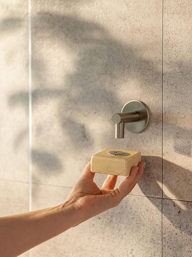 Hand greift zum festen Duschshampoo, das am Seifenhalter in der Dusche hängt