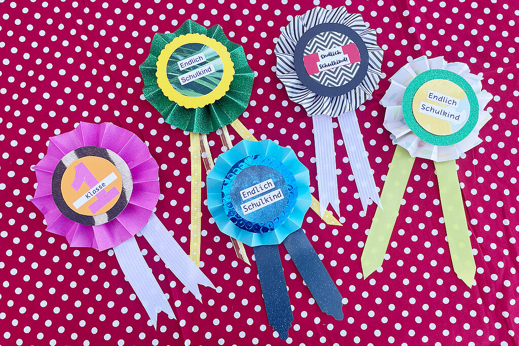 Einschulung Orden basteln mit bunten Papierrosetten und kreativen Schulkind-Motiven auf gepunktetem Hintergrund