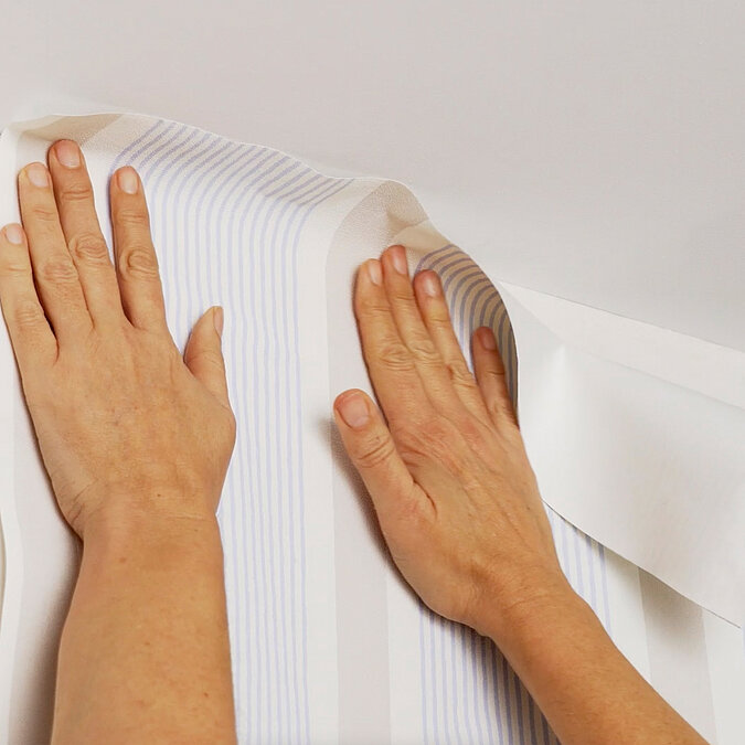 Wände richtig tapezieren: Tapetenbahn wird sorgfältig mit beiden Händen überlappend an der Wand angedrückt für saubere Übergänge und perfektes Tapezierergebnis
