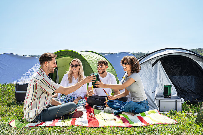 Festival Gadgets im Einsatz beim Camping mit Freunden vor Zelten, tragbarer Lautsprecher und Picknickdecke auf einem Festivalgelände