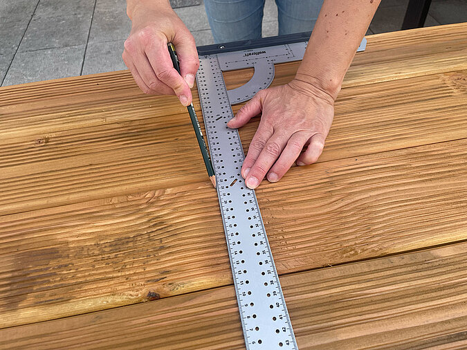 Grillplatz für die Terrasse bauen: Ablagebretter aus Holz werden mit Metallwinkel und Bleistift exakt angezeichnet – sorgfältige Vorbereitung für präzise Montage der DIY-Außenküche.