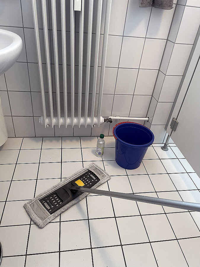 Fliesenboden wird mit Schrubber und Wischmopp geputzt daneben ein Putzeimer und eine Flasche Essigessenz