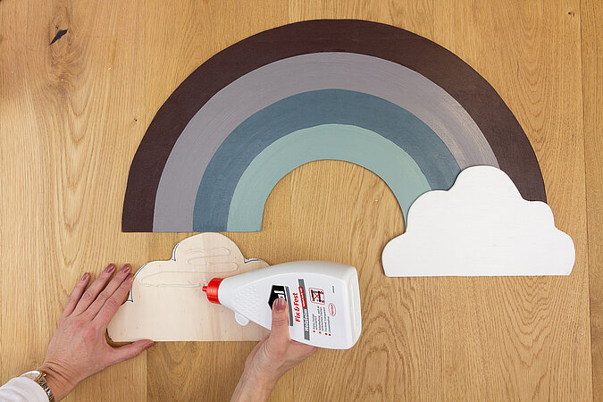 Wandleuchte im Regenbogen-Design bauen – Holz-Wolke wird mit Leim an den bemalten Regenbogen geklebt.
