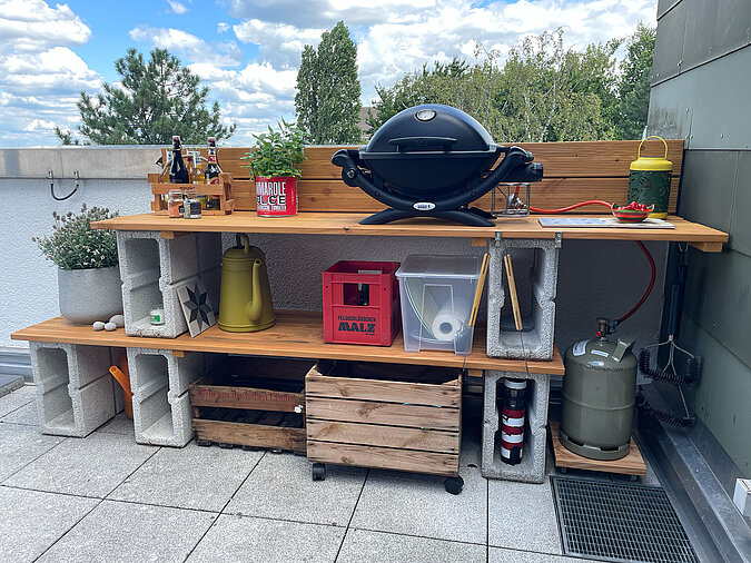 Grillplatz für die Terrasse bauen: DIY-Außenküche aus Betonsteinen und Holz mit Gasgrill, Gewürzen, Getränken und praktischen Aufbewahrungslösungen auf einer modernen Dachterrasse