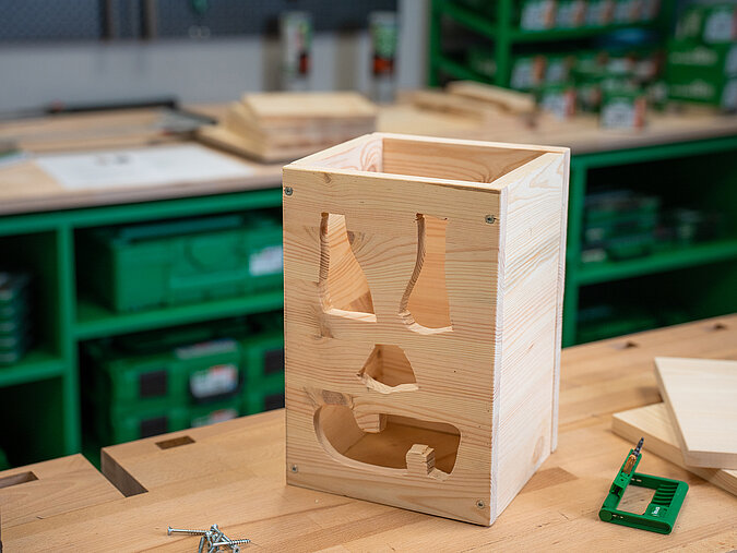Halloween-Laternen selber bauen: fertige, unbehandelte Holzlaterne mit ausgesägtem Gesichtsmotiv steht auf der Werkbank, bereit zum Dekorieren