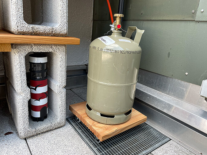 Grillplatz für die Terrasse bauen: Sicher platzierte Gasflasche auf Holzplatte mit Rollen neben DIY-Regal aus Betonsteinen – gut integrierte Techniklösung für den Außenbereich