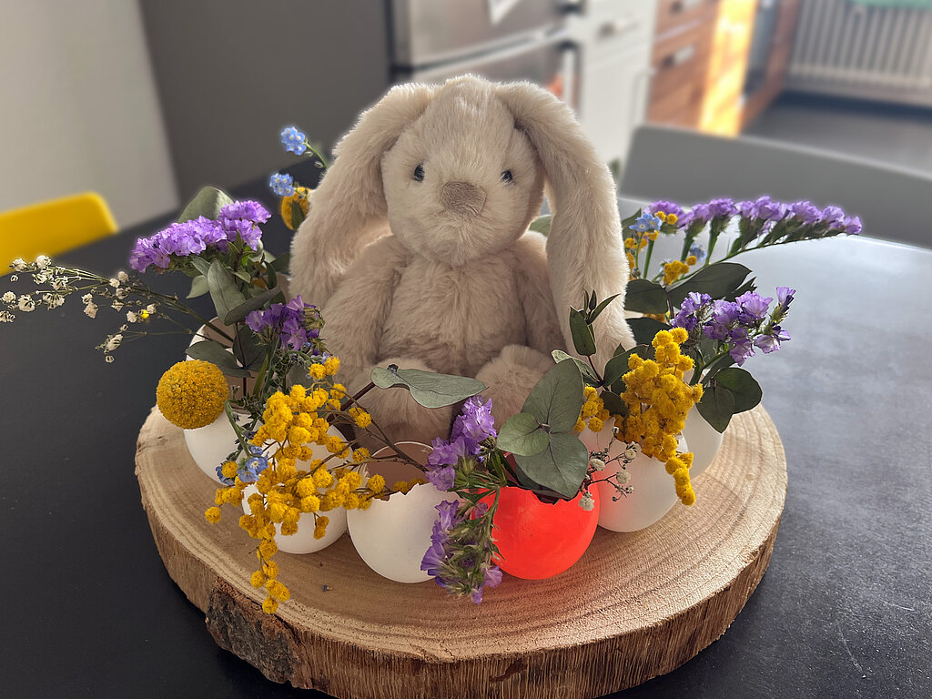 Osterkranz aus Eierschalen basteln als fertige Osterdeko mit Blumen, bunten Eierschalen und einem kleinen Stoffhasen auf einer Holzscheibe