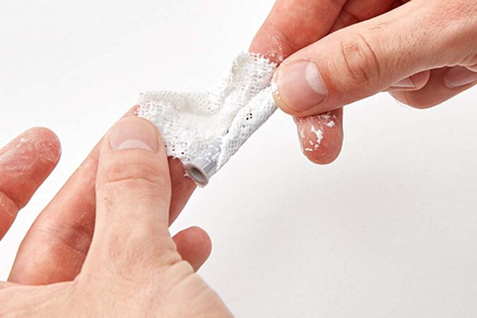 Zwei Hände halten einen Kunststoffdübel in der Hand, der Dübel wird mit dem befeuchteten Reparaturvlies umwickelt