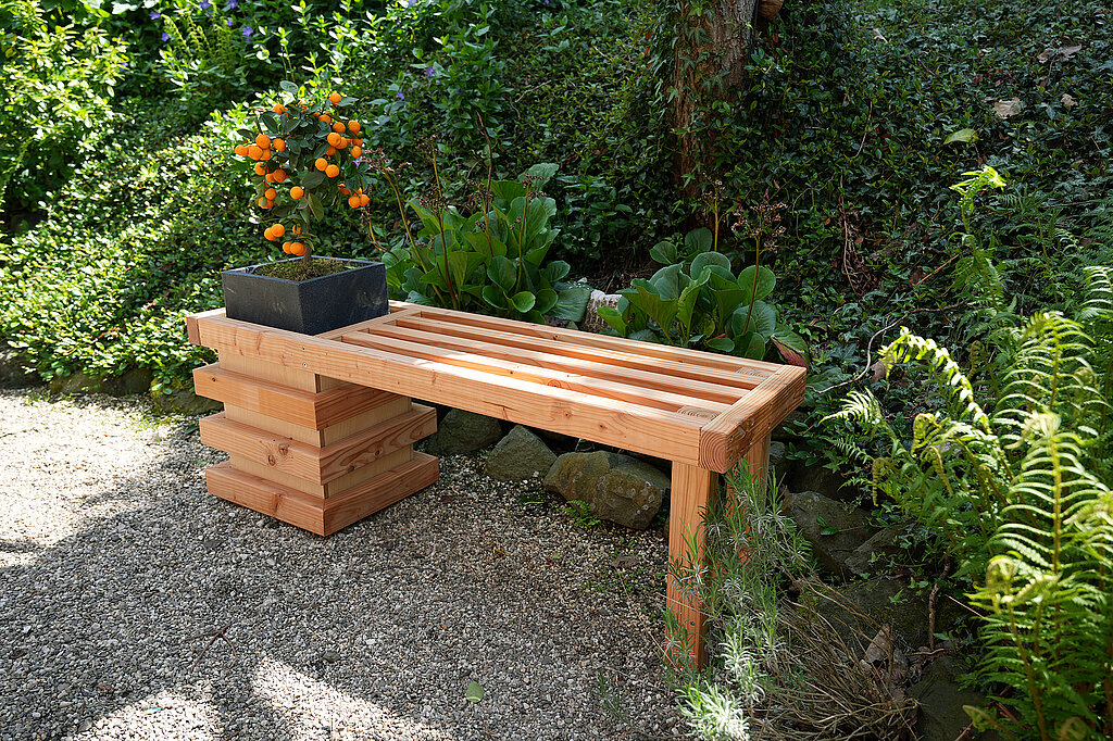 eine Gartenbank aus Holz mit an einer Seite integriertem Blumenkübel steht an einem schattigen Platz
