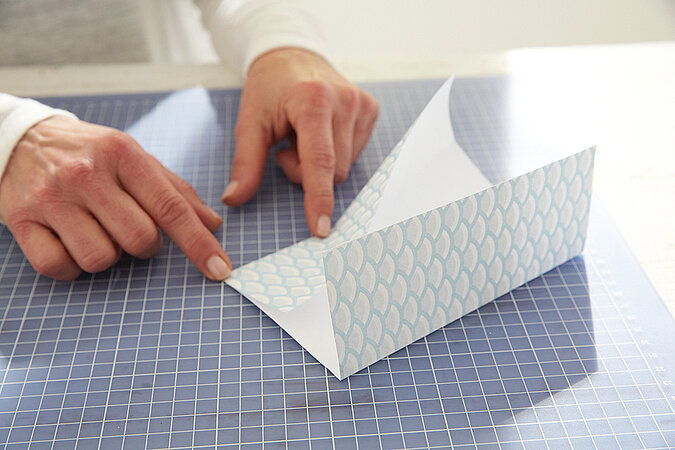 Hände falten ein hellblau gemustertes Papier zu einer Bestecktüte auf der Schneidematte
