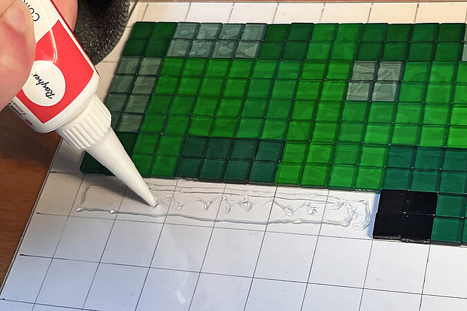 Creeper-Deko-Leuchte basteln mit Bastelkleber beim schrittweisen Befestigen der Mosaiksteine auf der Papier-Rastervorlage
