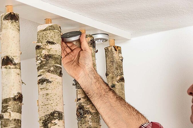 Batteriebetriebenes Licht wird zwischen Decke und Birkenstamm am Raumteiler aus Birkenstämmen angebracht