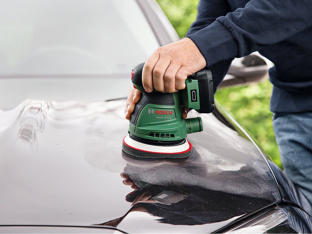Auto selbst waschen und polieren – Hand hält Poliermaschine auf Motorhaube, um Lack nach der Reinigung zu versiegeln und Hochglanz zu erzielen.