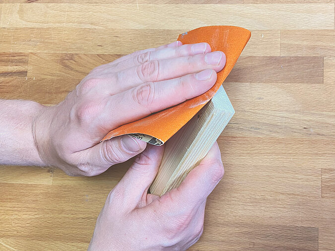 Eine Hand schleift mit Schleifpapier die Kanten eines Holzstücks – Vorbereitung für das DIY-Podest für die LED-Leute aus Getränkedose.