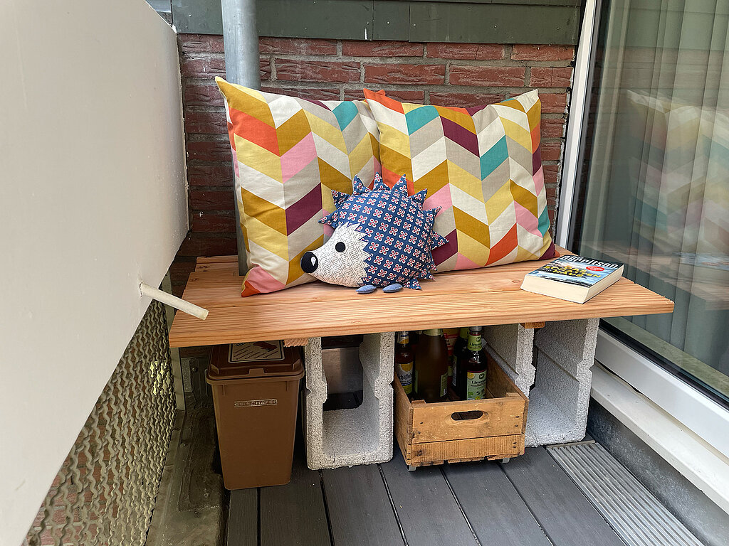 Aus Terrassendielen und Mauersteinen wurde eine gemütliche Sitzecke mit bunten Kissen und Stauraum gebaut – ideal für kleine Balkone oder Terrassen mit wenig Platz.