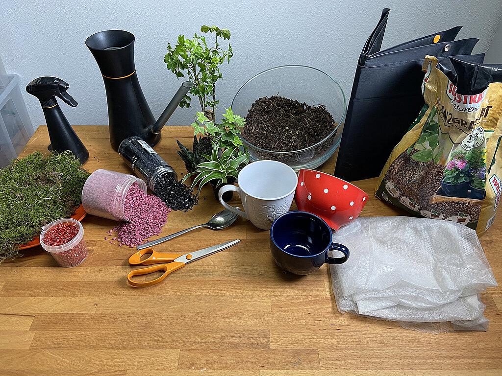 Tassengarten basteln mit umfangreichen Materialien wie Moos, Deko-Steinen, Pflanzen, Erde, Tassen, Werkzeug und Granulat auf einem Holztisch