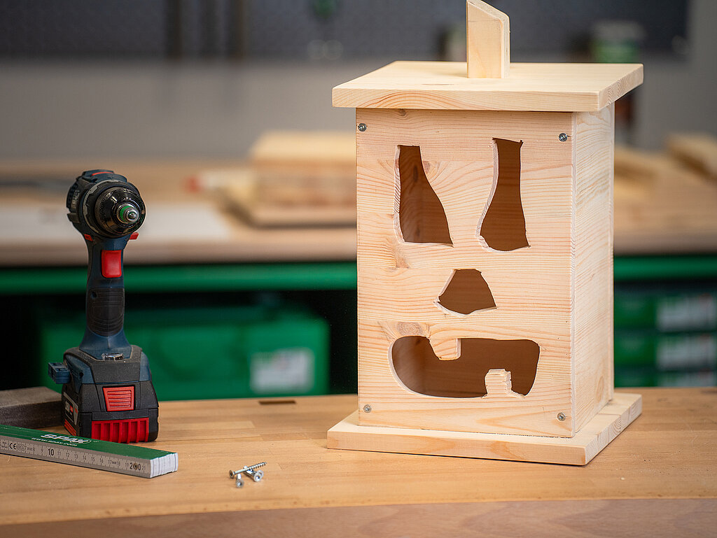 Halloween-Laternen selber bauen: unbehandelte Holzlaterne mit ausgesägtem Gesichtsmotiv auf einer Werkbank, daneben ein Akkuschrauber – Bastelprojekt in der Entstehung