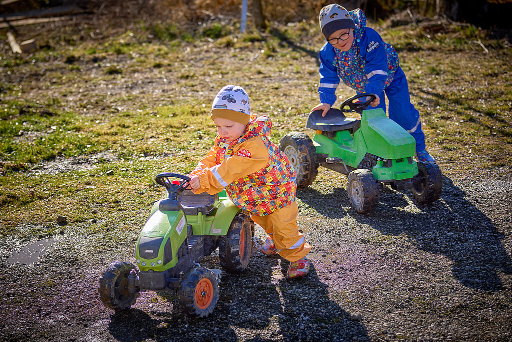 Tipps zum Heimwerken mit Kindern: Zwei kleine Kinder schieben ihre Trettraktoren über einen matschigen Untergrund – spielerisches Lernen von Einsatz und Bewegung beim Outdoor-DIY.