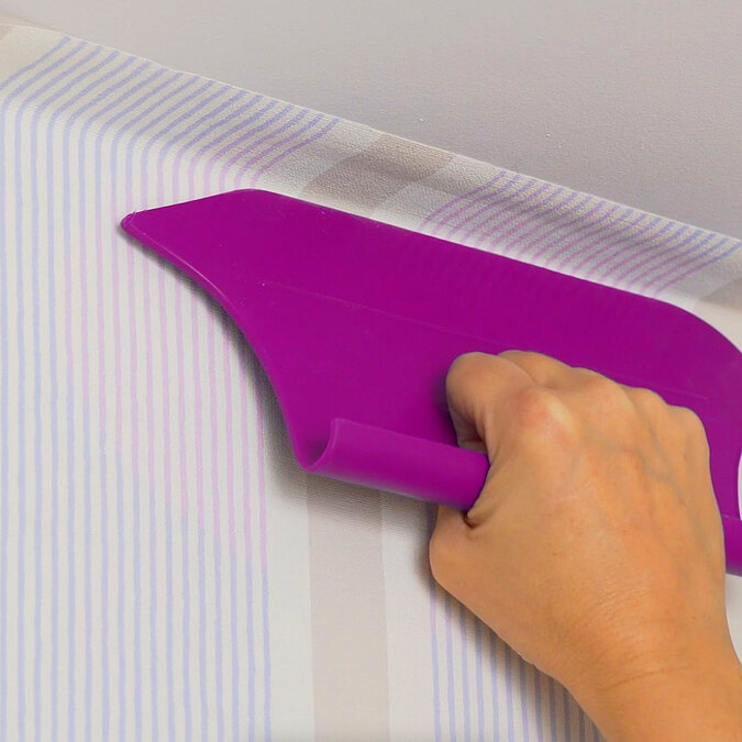 Wände richtig tapezieren: Tapetenbahn wird sorgfältig mit einem Spachtel überlappend an der Wand angedrückt für saubere Übergänge und perfektes Tapezierergebnis
