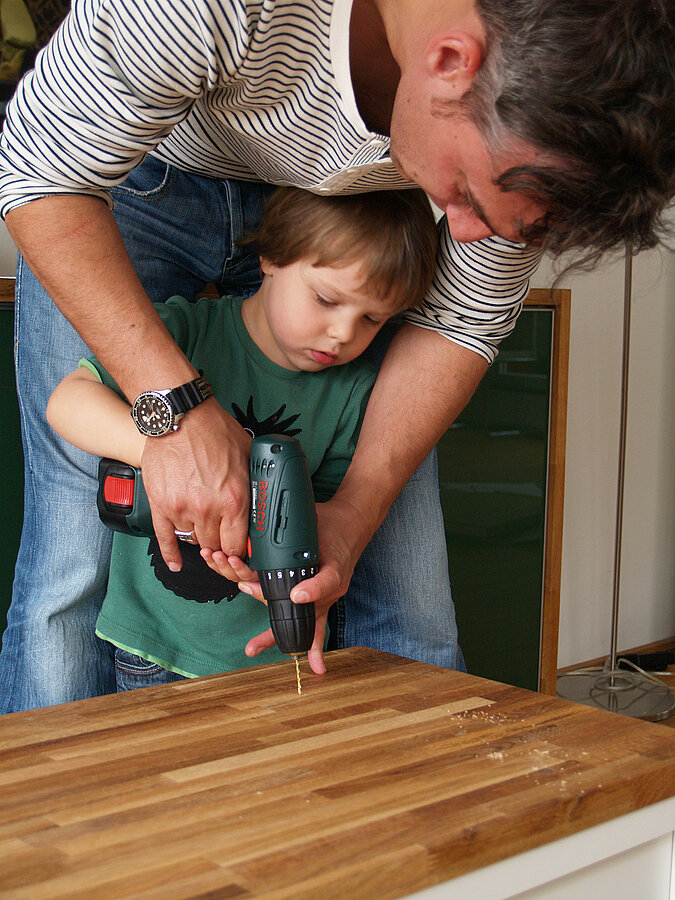 Tipps zum Heimwerken mit Kindern: Vater unterstützt seinen Sohn beim sicheren Umgang mit der Bohrmaschine – gemeinsames Schrauben als lehrreiches DIY-Erlebnis für Kinder.