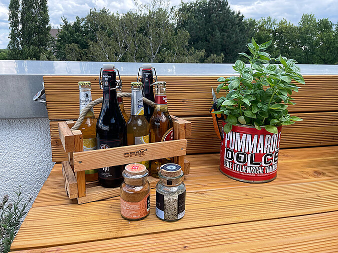 Grillplatz für die Terrasse bauen: Detailaufnahme einer DIY-Grillstation mit Bierflaschen im rustikalen Holzträger, frischem Basilikum im upgecycelten Tomatendosen-Topf und Grillgewürzen auf einer massiven Holzarbeitsfläche