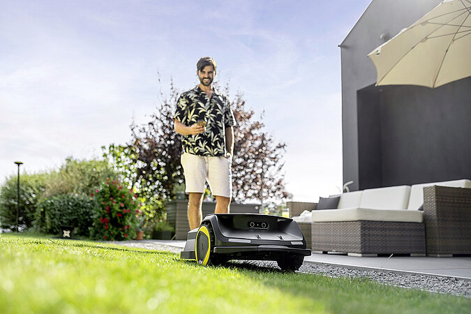 junger Mann in Shorts steuert Maehroboter per Smartphone